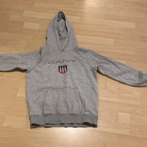 Grå hoodie från GANT - Säljer en stilren grå hoodie från GANT med deras ikoniska logga och ett broderat emblem på bröstet. Tröjan har en klassisk känguruficka och en bekväm huva. Perfekt för en avslappnad stil.