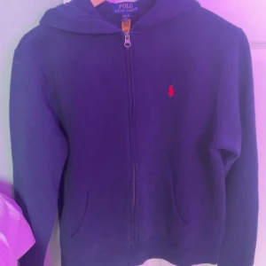 Marinblå hoodie med dragkedja från Ralph Lauren - Säljer en marinblå hoodie från Polo Ralph Lauren med dragkedja framtill och huva. Tröjan har en liten röd logga broderad på bröstet och två fickor fram. Perfekt för en avslappnad stil.