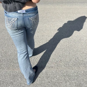 Lågmidjade bootcut jeans med paljettfickor - Säljer ett par ljusblå lågmidjade jeans med bootcut passform. Bakfickorna är dekorerade med paljetter och broderier i silver, vilket ger en unik och glittrig look. Perfekta för dig som vill sticka ut med dina jeans!
