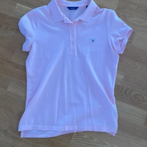 Ljusrosa pikétröja från GANT - Säljer en ljusrosa pikétröja från GANT med klassisk krage och knappar framtill. Tröjan har korta ärmar och liten broderad logga på bröstet. Perfekt för en stilren och sportig look!