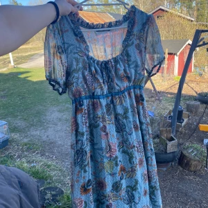 Blommig klänning med korta ärmar - Charmig blommig klänning i blått med korta ärmar och rund hals. Klänningen har ett färgglatt mönster med blommor i olika nyanser och en lätt rynkad detalj vid bysten. Står ingen storlek men skulle säga s/m
