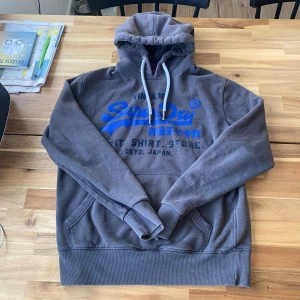 Grå hoodie från Superdry med blå logga - Säljer en grå hoodie från Superdry med stor blå logga och text framtill. Tröjan har huva med vita snören och känguruficka. Tillverkad i bomull och polyester, perfekt för en avslappnad stil. Passar L och M