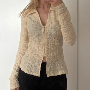 Beige texturerad topp från Gina Tricot - Säljer en beige texturerad topp från Gina Tricot med krage och knappar framtill. Toppen har långa ärmar och en unik textur som ger en stilren look. Den är aldrig använd eller testad då jag råkade beställa två likadana, så säljer den som finns kvar i original förpackning! 