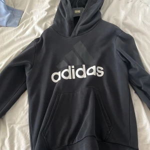 Svart hoodie från Adidas - Säljer en svart hoodie från Adidas med det klassiska logotyptrycket i vitt på framsidan. Tröjan har en stor ficka framtill och en bekväm huva. Perfekt för en avslappnad stil eller träning.