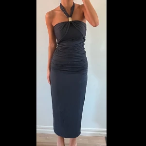 Mörkgrå fest klänning - Snygg mörkgrå festklänning med halterneck-design och dekorativt guldfärgat spänne. Klänningen har en tight passform med rynkade detaljer som ger en smickrande siluett. Perfekt för en kväll ute! Nyskick‼️