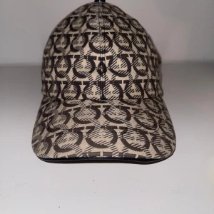 Beige och brun mönstrad keps från Ferragamo - Snygg keps från Ferragamo i beige och brunt med ett grafiskt mönster över hela ytan. Justerbar rem baktill för perfekt passform. Perfekt accessoar för en trendig look.