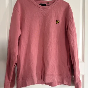 Rosa sweatshirt från Lyle & Scott - Säljer en rosa sweatshirt från Lyle & Scott med klassisk rund halsringning och broderad gul logga på bröstet. Tröjan har långa ärmar och ribbade muddar vid ärmslut och nederkant. Perfekt för en avslappnad stil.