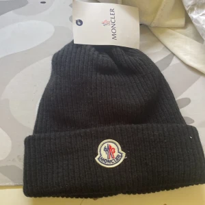 Svart moncler mössa - Svart ribbstickad mössa från Moncler med uppvikt kant och klassisk Moncler-logga framtill. Tillverkad i mjukt material som håller värmen och ger en stilren look. Perfekt accessoar för kyliga dagar.