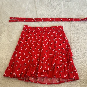 Röd blommig kjol med volang - Charmig röd kjol med små vita blommor och volangdetaljer. Kjolen har en omlott-design och ett matchande knytband i midjan. Perfekt för en somrig look!