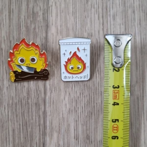 Roliga pins med eldtema, calcifer levande slottet - Två färgglada pins med eldtema, calcifer frånndrt levande slottet i ghiblis studios film. Den ena föreställer en glad eld på en stock, och den andra en eld i en papperskorg med japansk text. Perfekta för att piffa upp en jacka eller väska!