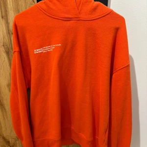 Orange hoodie från Amisu - Säljer en klarorange hoodie från Amisu i storlek M. Tröjan har huva och en liten vit textdetalj på bröstet. Perfekt för dig som vill sticka ut med färg. Mjuk och skön, passar till många tillfällen.