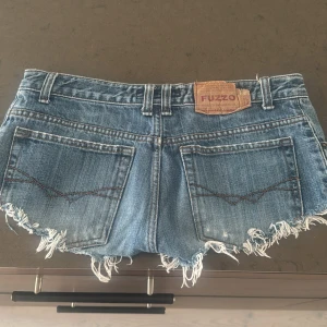Blå jeansshorts med fransar från Fuzzo - Säljer ett par blå jeansshorts från Fuzzo med slitna detaljer och fransig kant. Shortsen har klassiska fickor både fram och bak samt bälteshällor. Perfekta för en avslappnad stil.