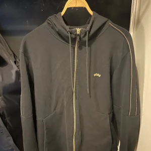 Svart hoodie från Boss - Säljer en stilren svart hoodie från Boss med dragkedja och huva. Tröjan har långa ärmar och dekorativa sömmar i kontrastfärg. Perfekt för en avslappnad stil.