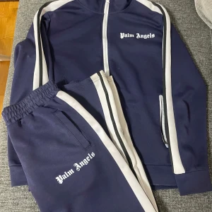 Mörkblå träningsjacka från Palm Angels - Säljer en snygg mörkblå träningsjacka från Palm Angels med vita detaljer. Använd ett par gånger inga större defekter med dressen, byxorna ingår, vid frågor skicka ett pm