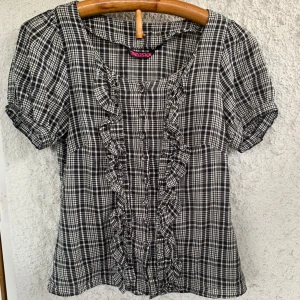 Svartvit rutig blus med volang från y2k - Supersöt svartvit rutig vintage blus från Indiska med korta puffärmar och volangdetaljer längs knappraden. Blusen är figursydd och  har klassisk krage och knappar framtill. Perfekt för dig som gillar en feminin och trendig stil.