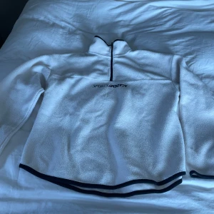 Vit fleece tröja  - Mysig vit fleece tröja med halv dragkedja och svarta detaljer vid krage och nederkant. Perfekt för kyliga dagar och sportiga aktiviteter.  