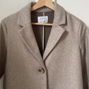 Beige kappa från Zara - Snygg beige kappa från Zara med klassisk design. Den har långa ärmar och stängs med två knappar framtill. Perfekt för höst och vår med sin stilrena look.