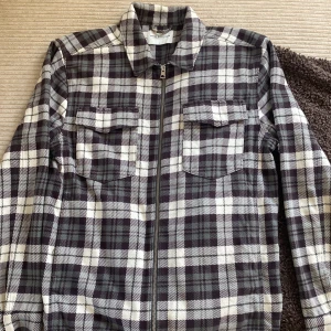 Rutig overshirt från These Glory Days - Säljer en snygg rutig overshirt från These Glory Days i svart, grått och vitt. Jackan har dragkedja framtill, två bröstfickor med lock och klassisk krage. Perfekt att slänga över en t-shirt för en avslappnad stil.