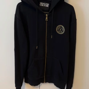 Svart hoodie från Versace Jeans Couture - Svart hoodie med dragkedja från Versace Jeans Couture. Stor logga i guld på ryggen och mindre logga på bröstet. Huva med dragsko och två fickor framtill. Perfekt för en snygg och avslappnad stil.