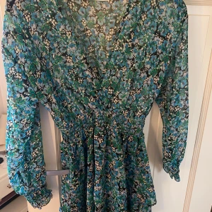 Blommig klänning från Zara - Säljer en vacker blommig klänning från Zara i gröna och blå toner. Klänningen har en v-ringad design med långa ärmar och ett smickrande snitt i midjan. Perfekt för vår och sommar!