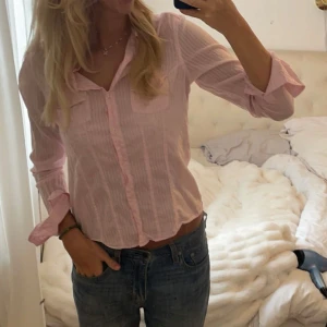 Rosa randig blus - Säljer en söt rosa blus med ränder och knappar framtill. Blusen har långa ärmar och en klassisk krage. Perfekt för en avslappnad stil.