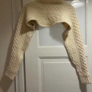 Beige stickad croppad polotröja - Supermysig croppad polotröja i beige med grovstickat flätmönster. Tröjan har långa ärmar och hög polokrage, perfekt för lager-på-lager. Den stickade strukturen ger en trendig och cozy vibe till din outfit.