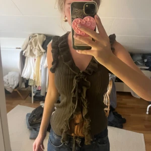 Olivgrön ribbad topp med volanger från Zara - Säljer en olivgrön ribbad topp från Zara med volanger längs axlar och framkant. Toppen har snörning framtill och är ärmlös med v-ringad hals.  Aldrig använd prislapp kvar! Superfin