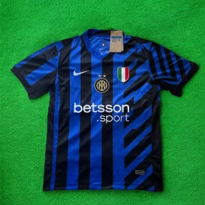 Inter Milan matchtröja Nike blå/svart - Inter Milan fotbollströja från Nike i blått och svart med coola ränder och guldiga detaljer. Tröjan har klubbmärke, sponsortryck och italienska mästarmärket på bröstet. Tillverkad i lätt och ventilerande material, perfekt för match eller träning.