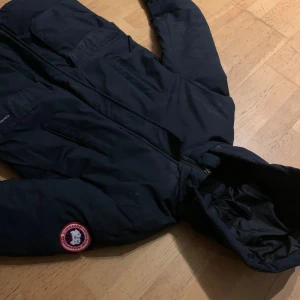 Canada goose jacka - Säljer en svart dunjacka från Canada Goose i bra skick. Jätte varm och skön nu inför vintern. Jackan är inte äkta och har ett litet hål vid fickan. Saknar även 2 knappar vid dragkedjan