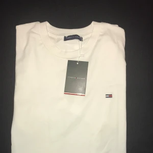 Vit t-shirt från Tommy Hilfiger - Vit t-shirt från Tommy Hilfiger med klassisk rund hals och korta ärmar. Diskret Hilfiger-logga broderad på bröstet och mjukt bomullsmaterial som känns skönt mot huden. Perfekt basic med clean look och snygg passform.