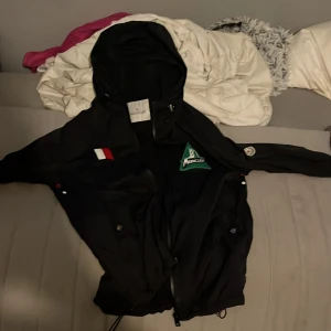 Svart vindjacka från Moncler med huva - Svart vindjacka från Moncler med huva och dragkedja. Jackan har coola patchar på bröstet och ärmen, samt snörning i nederkant. Perfekt för dig som vill ha en stilren och sportig look. Materialet är lätt och känns smidigt.