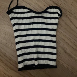 Randig ribbad tubtopp i vitt och svart - Snygg ribbad tubtopp med svarta och vita horisontella ränder. Toppen är ärmlös och har en bandeau-form med smala axelband. Perfekt för en trendig och avslappnad look under varma dagar.