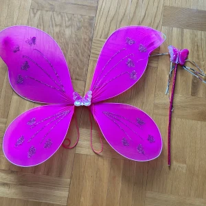 Rosa fjärilsdiadem med vingar - Säljer ett set med ett rosa diadem med fjäril och matchande stora fjärilsvingar. Diademet har glittriga detaljer och silvriga band, vingarna är dekorerade med glitterfjärilar och har elastiska band för att sitta bra. Perfekt för maskerad eller temafest.