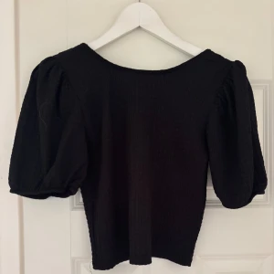 Svart topp med puffärmar - En svart topp med snygga puffärmar och rund halsringning. Tyget är lätt ribbat och ger en cool struktur. Perfekt för dig som gillar en stilren men trendig look. Passformen är normal och toppen är lite croppad.