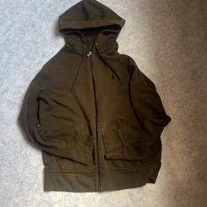 Mörkgrön hoodie från Polo Ralph Lauren - Säljer en mörkgrön hoodie från Polo Ralph Lauren med dragkedja och snörning vid huvan. Tröjan har långa ärmar, två fickor fram och en liten broderad logga på bröstet. Perfekt för chill dagar och enkel att matcha med jeans eller joggers.