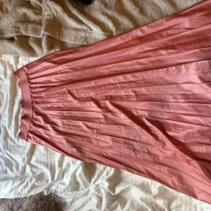 Plisserad rosa långkjol från VILA - Säljer en snygg plisserad långkjol från VILA i en mjuk rosa färg. Kjolen har ett brett, elastiskt midjeband och är gjord i ett lätt, glansigt syntetmaterial som faller fint. Perfekt för dig som gillar feminina och trendiga plagg.