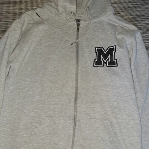 Grå hoodie med M från Mira Paris - Snygg grå hoodie från Mira Paris i ekologisk bomull. Framtill finns ett stort svart M-tryck, dragkedja och två fickor. Huva med snörning och mjukt material som känns chill. Perfekt för dig som gillar enkel stil med en twist.