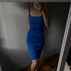 Blå tight klänning - Säljer en snygg blå klänning med tight passform och ärmlös design. Klänningen har breda axelband, fyrkantig urringning och en liten slits bak. Materialet är mjukt och stretchigt, perfekt för dig som gillar en enkel men stilren look.