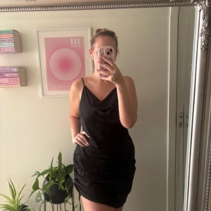 Svart omlottklänning från Amisu - Säljer en svart kort klänning från Amisu med tunna axelband och snygg draperad omlottdetalj framtill. Klänningen har snörning på sidan för att justera passformen och är gjord i ett mjukt, glansigt polyestertyg som ger en lyxig känsla. Väldigt strechig och passar en L-XL. Man kan styrka längden på klänningen med hjälp av banden på sidan 