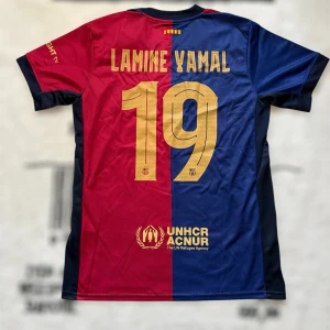 FC Barcelona Lamine Yamal tröja Nike S - FC Barcelona matchtröja från Nike med Lamine Yamal och nummer 19 på ryggen. Tröjan är kortärmad och har klassiska blå och röda paneler med guldiga detaljer, klubbmärke och sponsorer. Tillverkad i lätt och ventilerande polyester, perfekt för fotbollsälskare.
