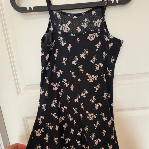 Svart blommig kortklänning med smala axelband - Supersöt svart kortklänning med små blommor i rosa blått och vitt. Klänningen har smala axelband och lätt utställd passform. Materialet är mjukt och har en lätt struktur. Perfekt för sommarens alla tillfällen!