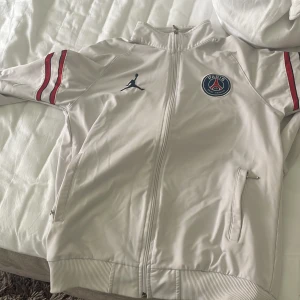 PSG x Jordan vit vindjacka - Snygg vit PSG x Jordan vindjacka med dragkedja, ribbade muddar och röda ränder på ärmarna. Jackan har broderad Jumpman-logga och PSG-märke på bröstet. Perfekt för dig som gillar sportig och clean stil.