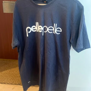 Blå t-shirt från Pelle Pelle - Snygg marinblå t-shirt från Pelle Pelle med vit logga tryckt på bröstet och mindre logga i nacken. Klassisk rund halsringning och normal passform. Perfekt för dig som gillar streetwear och enkel stil.