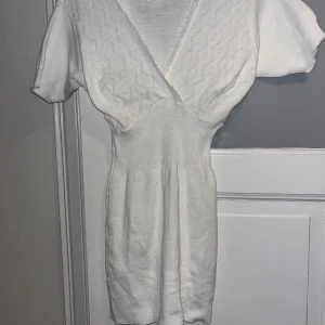 Vit stickad kortklänning med spets - Supersöt vit stickad kortklänning med v-ringad omlott upptill och korta ärmar. Klänningen har ett diskret spetsmönster på bröstet och ribbad midja som ger en snygg siluett. Perfekt för dig som gillar mjuka material och stilrena detaljer.