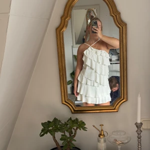 Vit volangklänning med smala axelband - Supersöt vit kortklänning med flera lager volanger och smala axelband som korsas framtill. Klänningen har en luftig passform och öppen rygg, perfekt för varma dagar. Materialet känns lätt och flowy, vilket ger en riktigt somrig vibe.