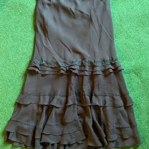 Brun volangkjol från KappAhl - Supersöt svart kjol från KappAhl med flera volanglager och spetsdetaljer. Kjolen har en mjuk, flowy känsla och är perfekt för dig som gillar romantisk stil. Den är knälång och har en feminin siluett som ger en snygg look.