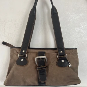 Brun axelväska från Esprit - Snygg brun axelväska från Esprit med svarta skinnhandtag och coolt bältedetalj framtill. Väskan har dragkedja upptill och flera praktiska fack inuti. Perfekt storlek för att få med allt du behöver och passar till många olika stilar.