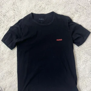 Svart t-shirt från Hugo Boss - Svart t-shirt från Hugo Boss med röd HUGO-logga på bröstet. Klassisk rund hals och korta ärmar. Tillverkad i mjuk bomull som känns skön mot huden. Enkel och stilren design som funkar till allt, perfekt för dig som gillar cleana plagg.