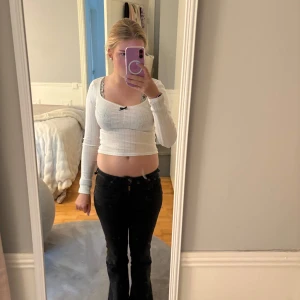 Vit croppad långärmad topp med rosett - Söt vit croppad topp med lång ärm och diskret hålmönster. Toppen har en liten svart rosett framtill och urringning. 