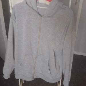 Grå hoodie med dragkedja och huva - En basic grå hoodie med dragkedja framtill och stor huva. Tröjan har två fickor på magen och långa ärmar. Perfekt för chill dagar eller när du vill ha något bekvämt och enkelt. Passar till det mesta och är lätt att matcha.